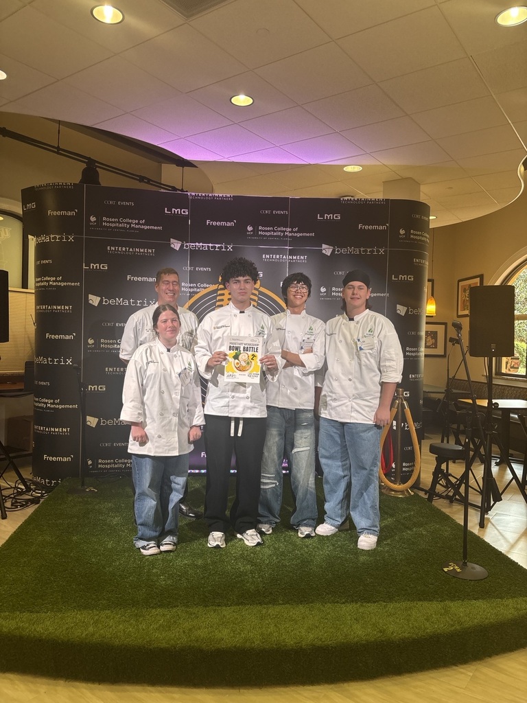 Five people in white coats and sneakers stand on a green platform in a room. A speaker is on the right. A black backdrop with text is behind them.