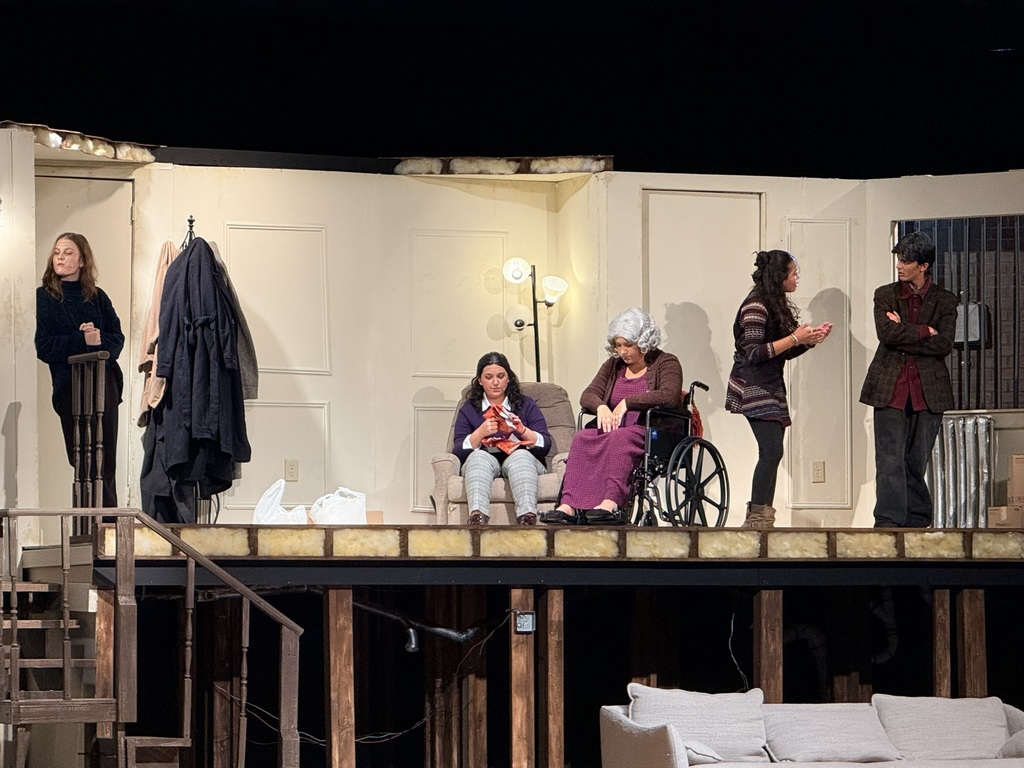 A group of people on stage with a wooden floor. One woman sits in a wheelchair. Two others hold coats.