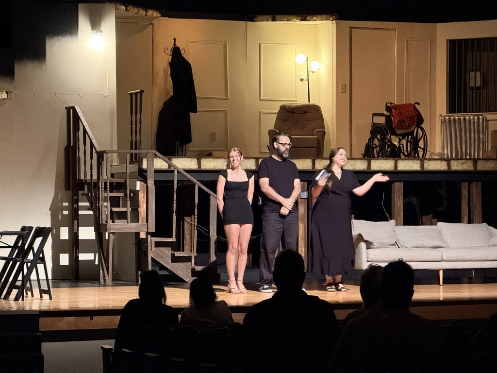 Four people stand on a stage. Two women wear black dresses. Two men wear black shirts. A couch is in the background.