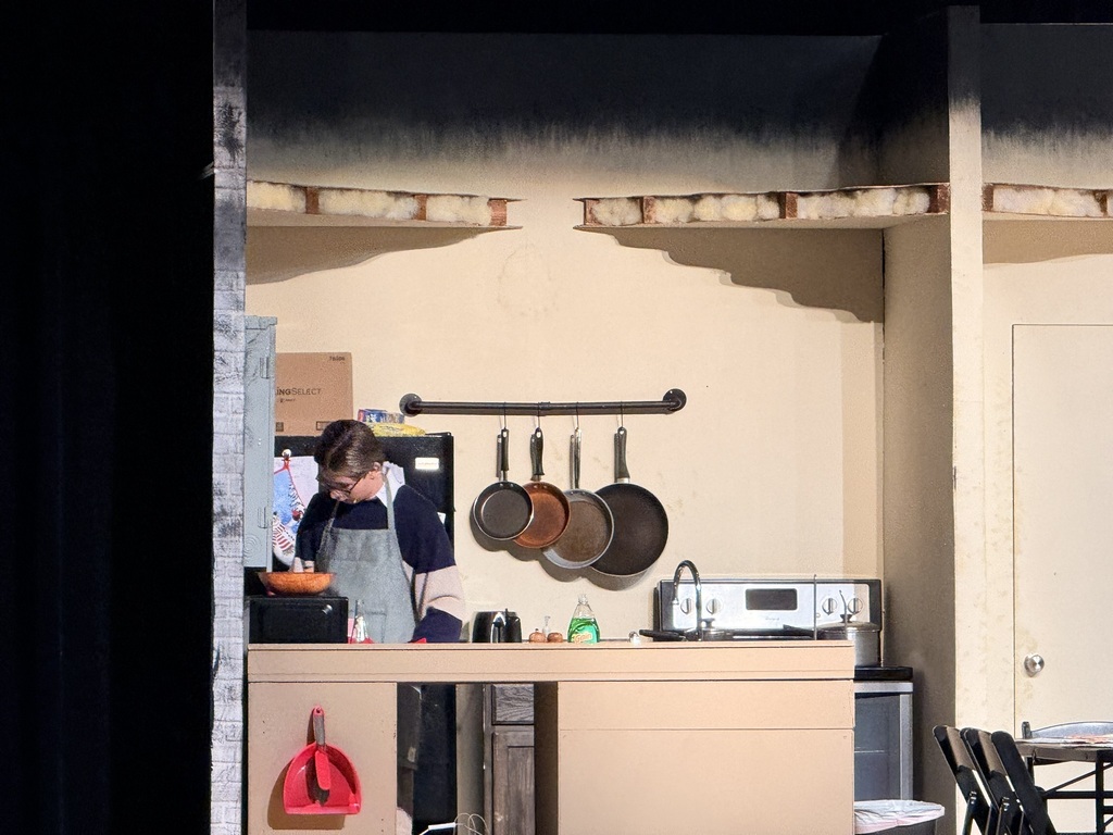 A kitchen scene with a person in apron, a microwave, and four hanging pots. The room has beige walls and an open door.