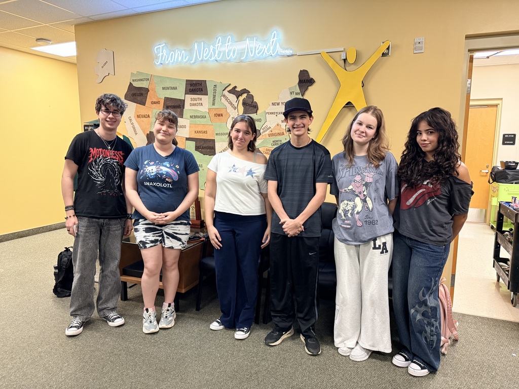 A group of seven young adults in casual clothing stand together in a brightly lit room with yellow walls and a map.