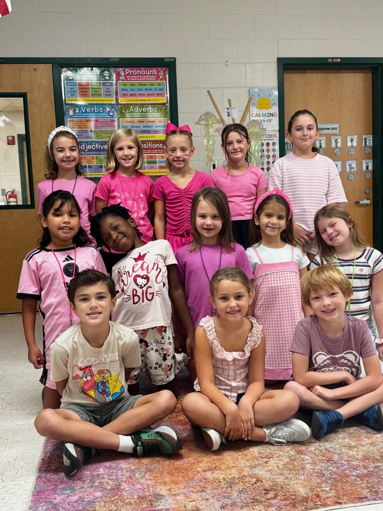 A group of young children in pink tops are seated and kneeling on a rug. Behind them, two doors are open.