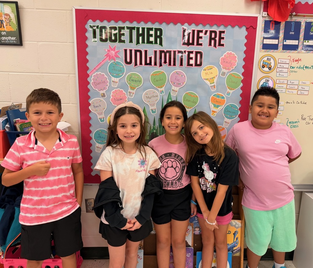 Five children, three girls and two boys, smiling in front of a classroom bulletin board.