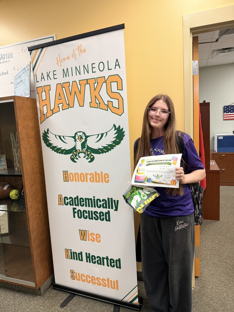 Person in purple shirt stands next to sign with "Lake Minneola Hawks" and "Honorable, Academically Focused, Wise, Kind Hearted, Successful" on a yellow background.