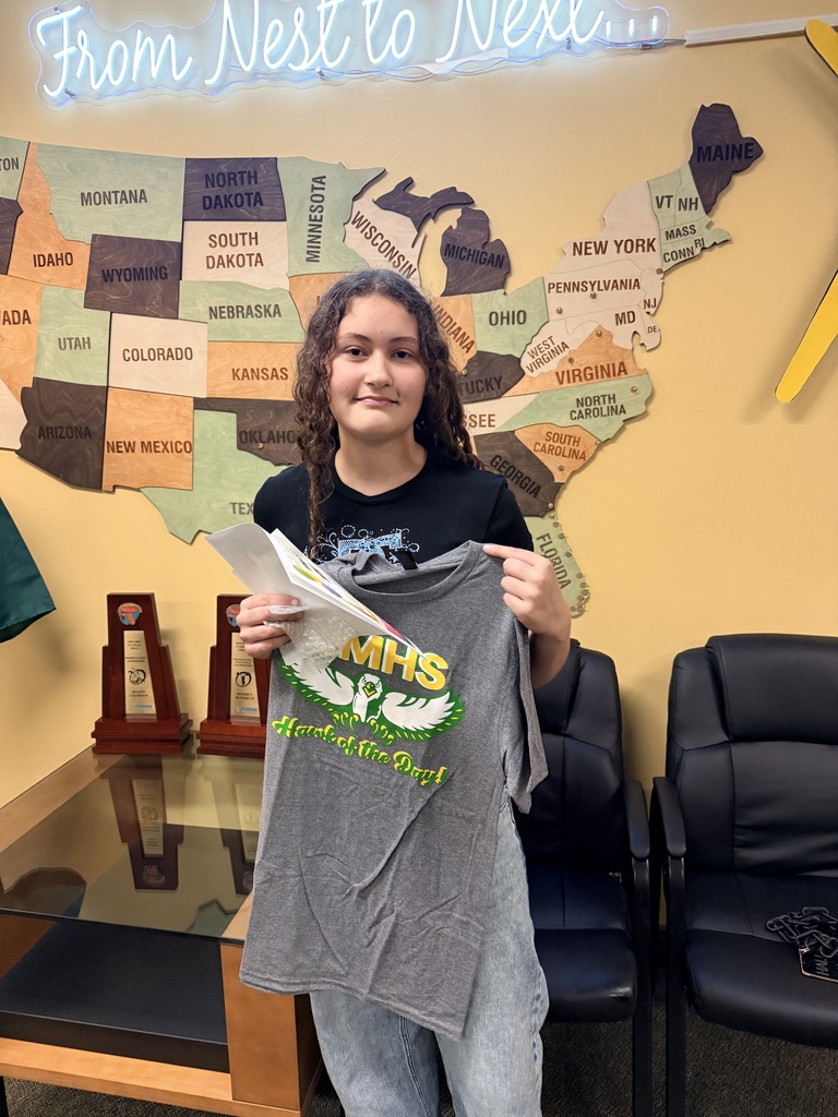 A young person holds a gray t-shirt with green text. They stand before a map of the US. A black chair is behind them.