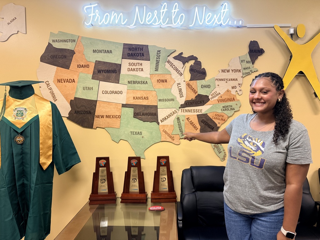 Woman in gray LSU shirt pointing to a map. Three trophies and a green graduation gown displayed on a glass table.