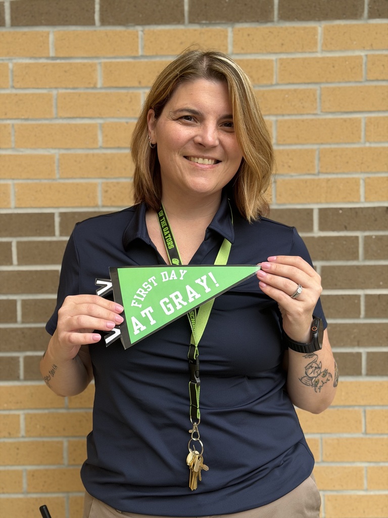 Woman in blue shirt holds a green pennant reading "First Day at Gray!" in front of brick wall.