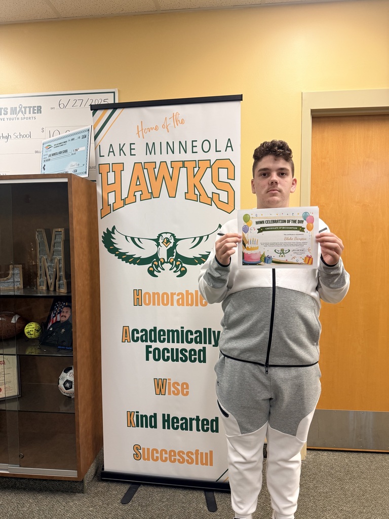 A man in a white and gray jacket holding a certificate stands in front of a banner that reads "Lake Minneola Hawks."
