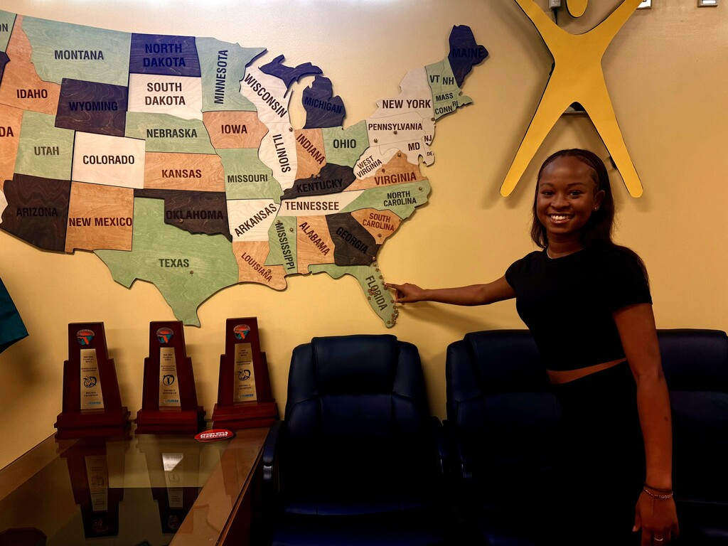 A woman stands by a map of the US, pointing at a state while seated in a blue chair.