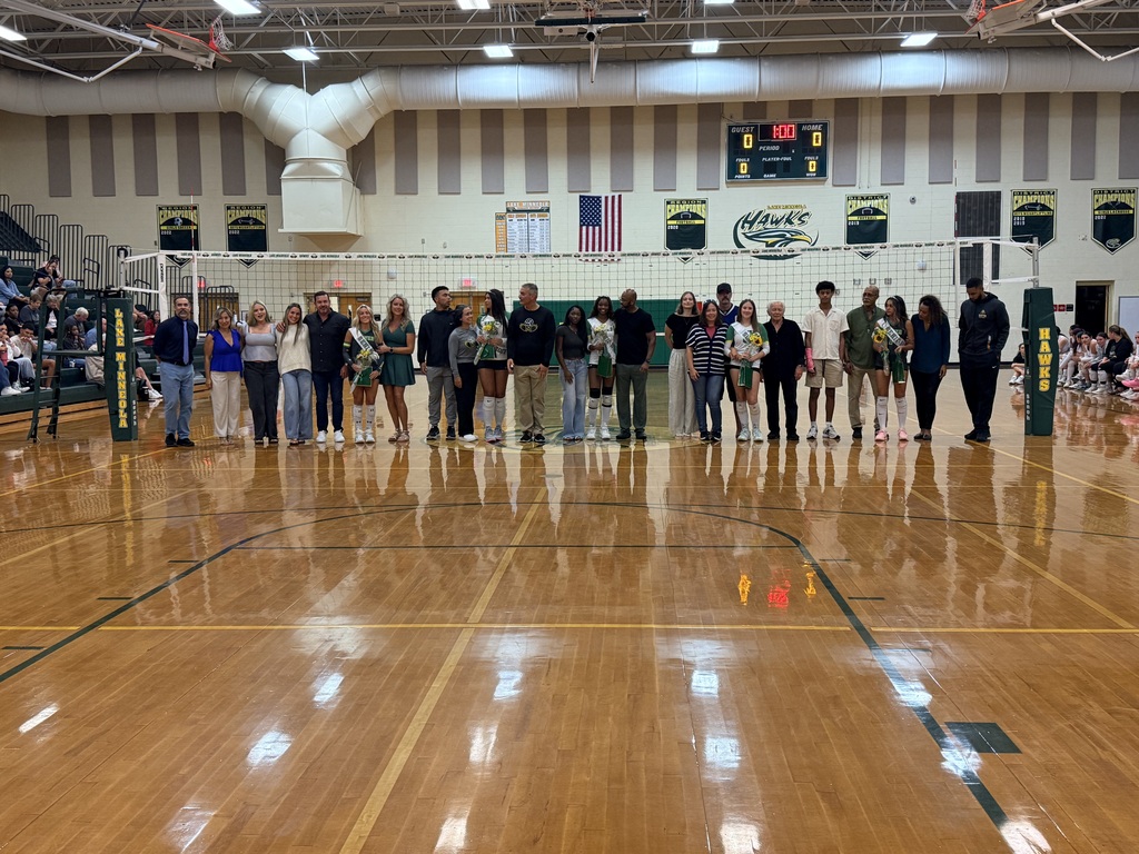 A gym with a volleyball net. A group of people stands in a row, facing the audience.