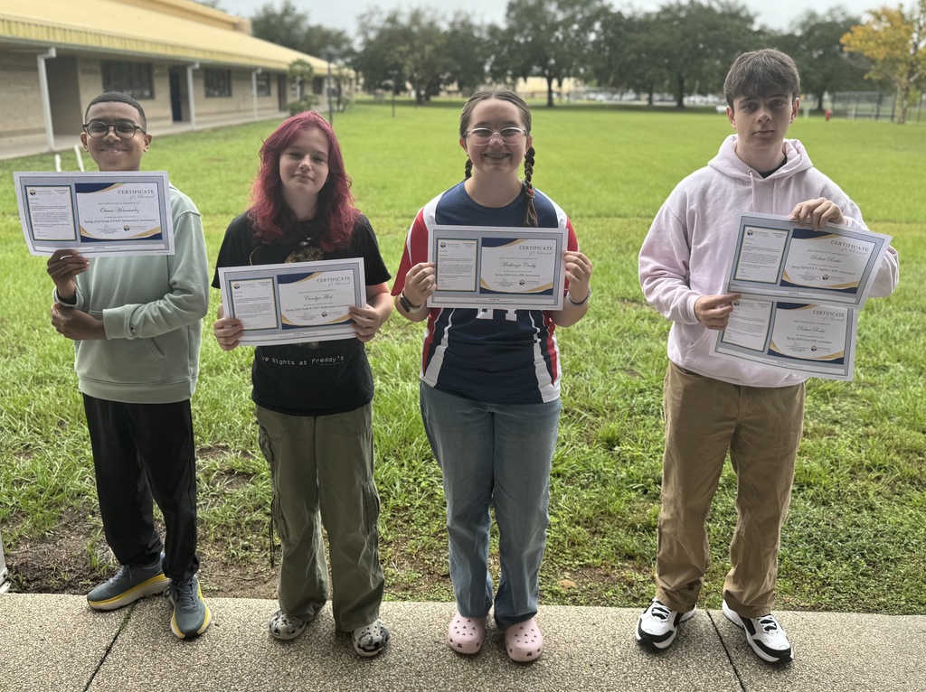 Four young people stand on a sidewalk with certificates. A grassy field and building are behind them.