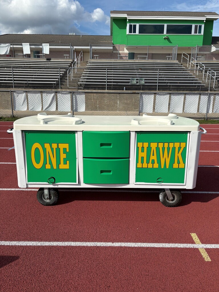 A rolling cart with green and white sections labeled "ONE" and "HAWK" on a track field with empty bleachers in the background.