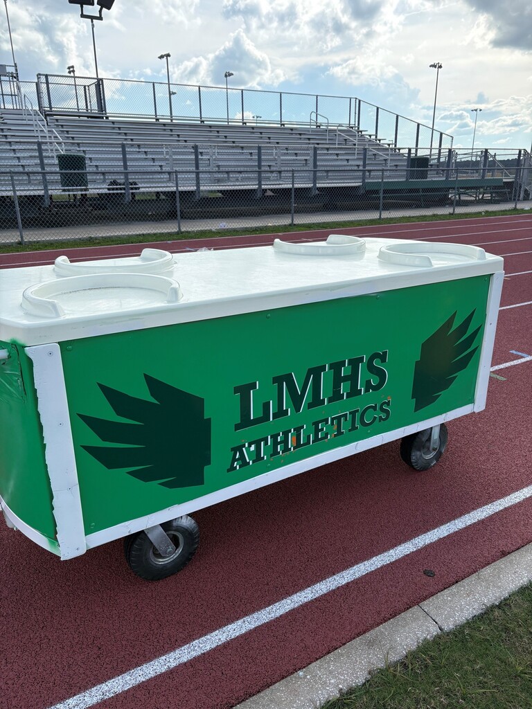 A green and white cart with "LMHS Athletics" printed on it on a track field with empty bleachers and a grassy area.