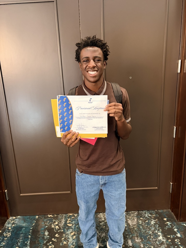 Man in brown shirt and jeans holding a certificate and smiling, standing in front of wooden doors.