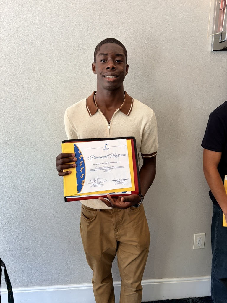 A man holds an award certificate and smiles. He wears a white shirt and brown pants. A person stands behind him, partially visible, next to a white wall.