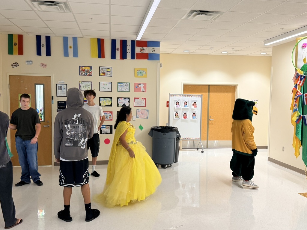 A classroom hallway with flags and decorations. Students in costumes, including one in a yellow dress, stand around a mascot.