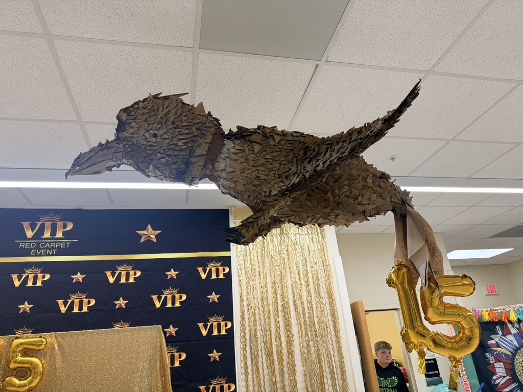 A hanging dinosaur piñata above a table. "VIP" and "15" signs on blue and gold banners. A person stands next to the table.