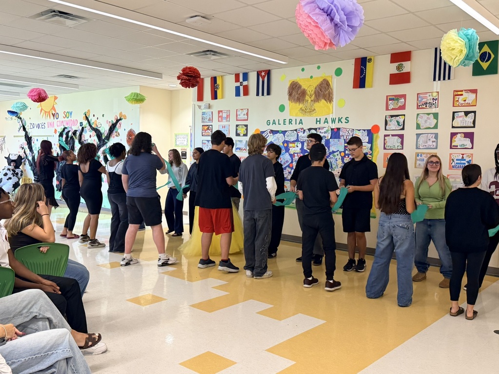 A group of people stand in a hallway with yellow floor tiles. Some are dancing while others sit on the floor. Flags and decorations adorn the walls.