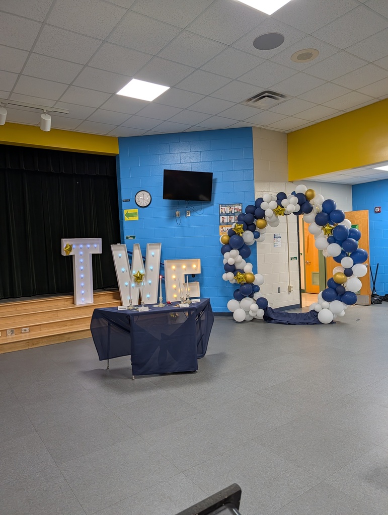 Room with blue walls and white ceiling. An empty stage with a black curtain is visible. A table displays 'TWE' letters.