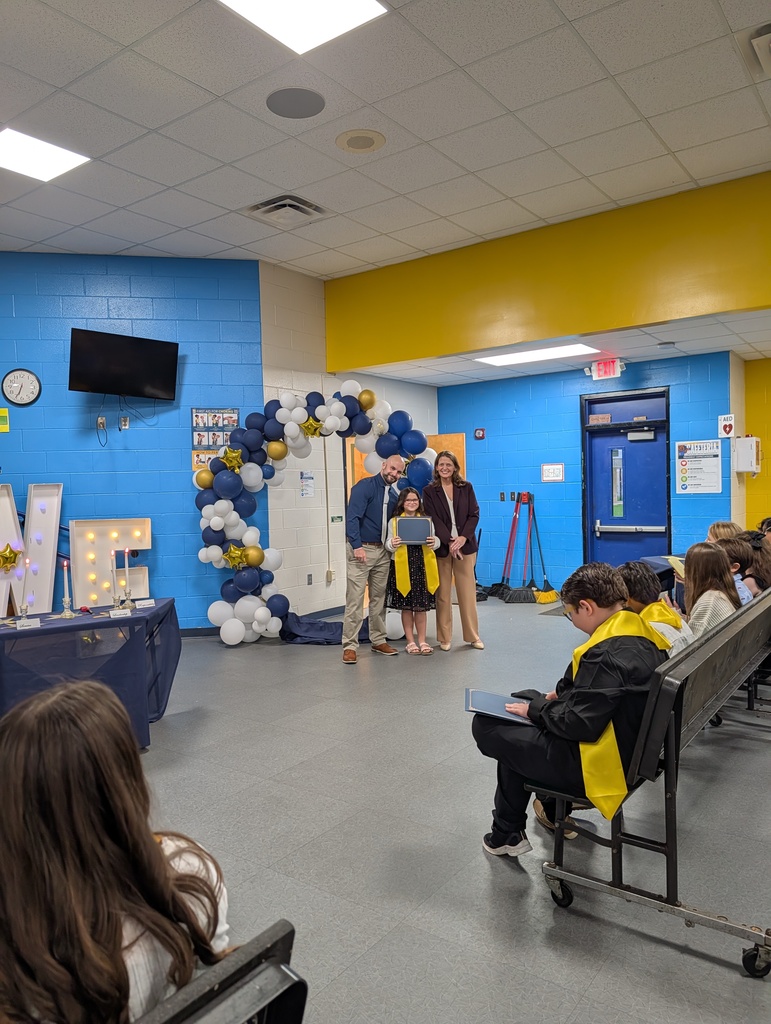 Several students and adults in a school hallway. A girl receives an award from two adults.
