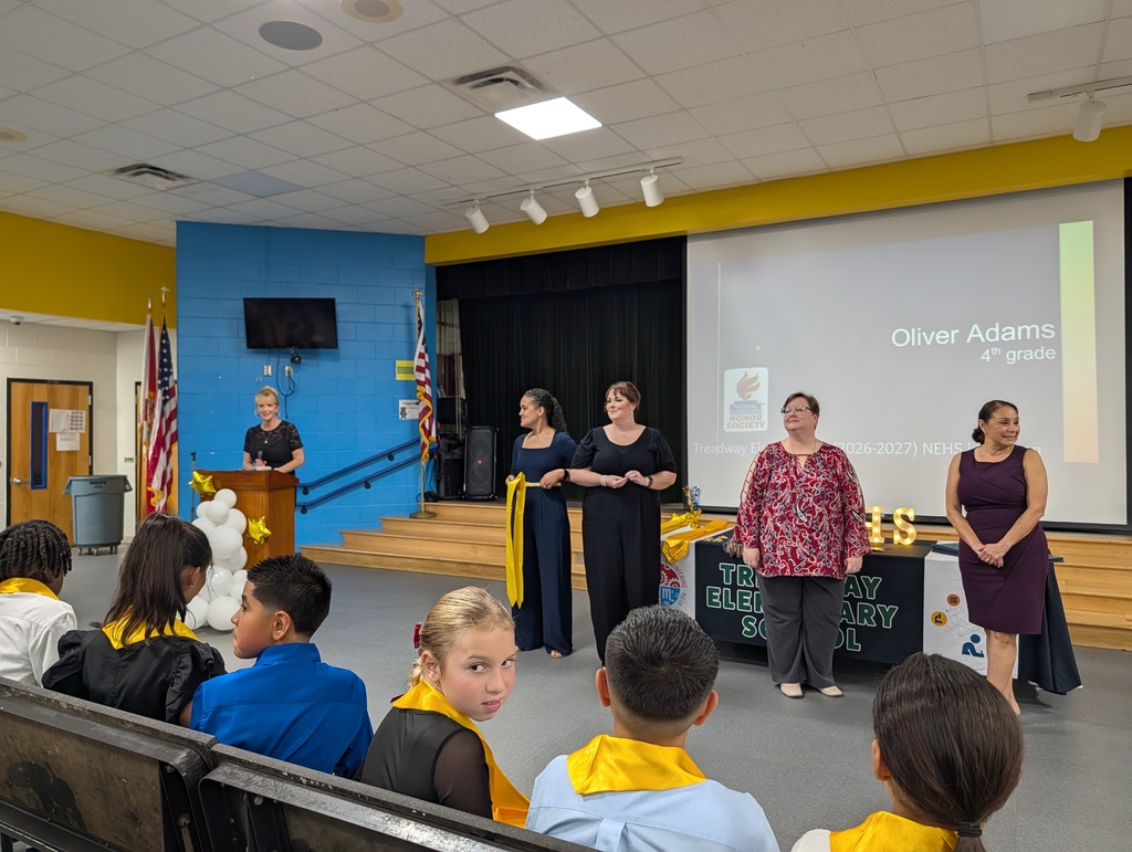 A group of adults stands on a stage with a presentation screen behind them. Children are seated in front.