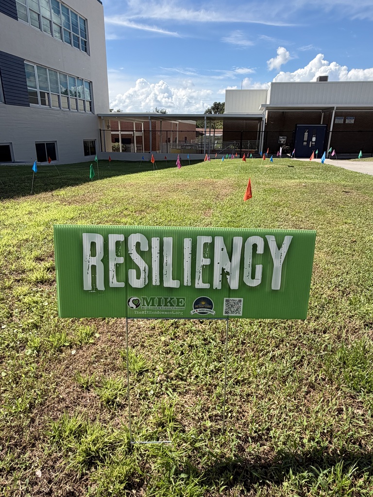 Green sign in grass reads "RESILIENCY" in white. Buildings with glass windows in background. Small orange cones and blue flags on lawn.