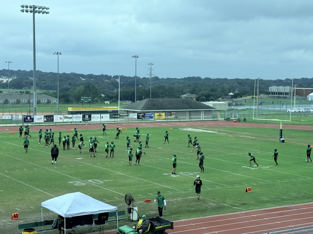 Football practice with players spread across field, some wearing white helmets. Tent and equipment on field side. Background shows stadium, trees, and buildings.