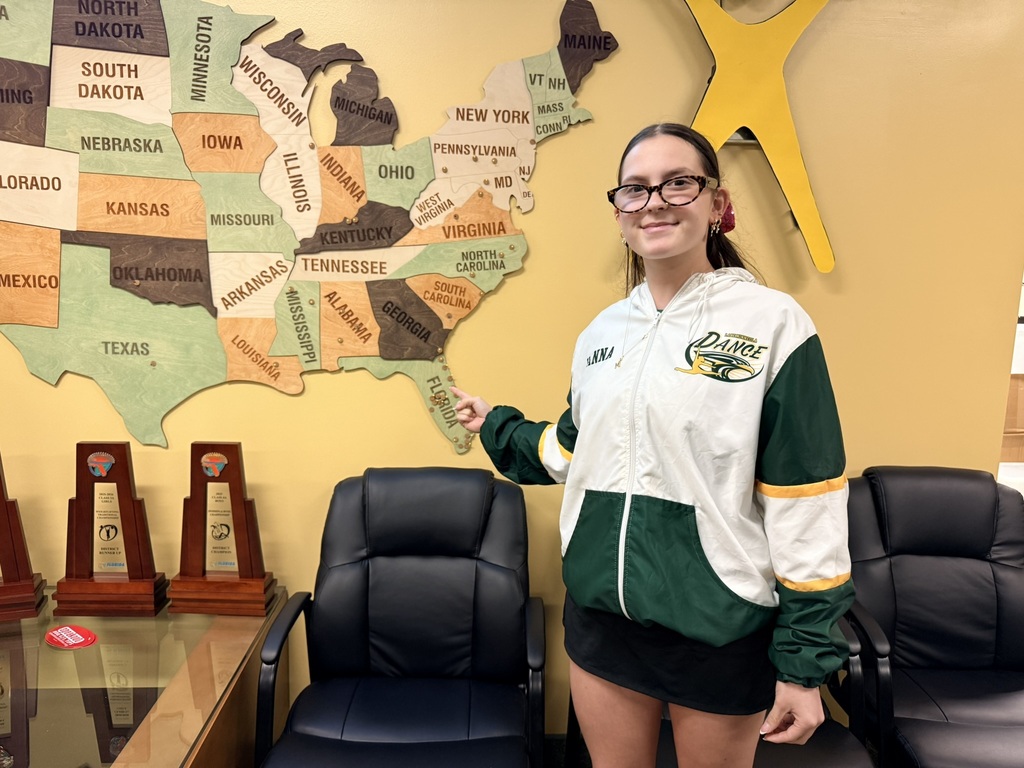 A girl in glasses and a sports jacket stands in front of a map of the U.S. and three trophies.