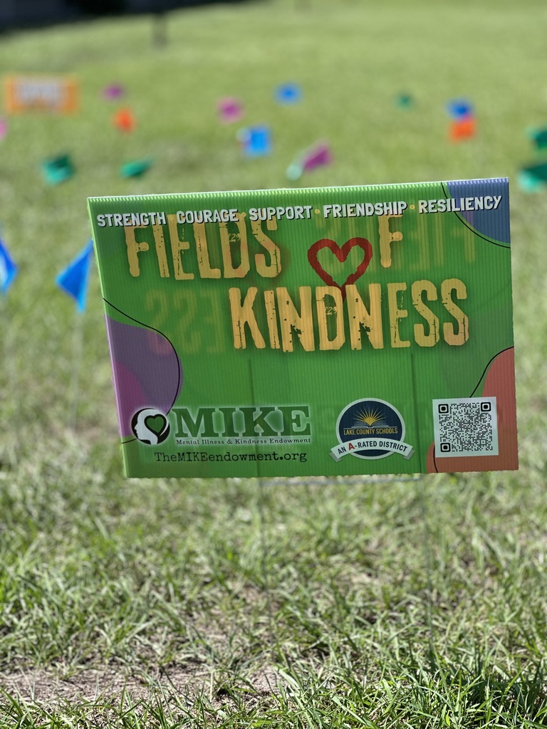 Green sign with white letters on grass. Sign says "Fields of Kindness" with a heart. Small flags are scattered on the grass behind it.
