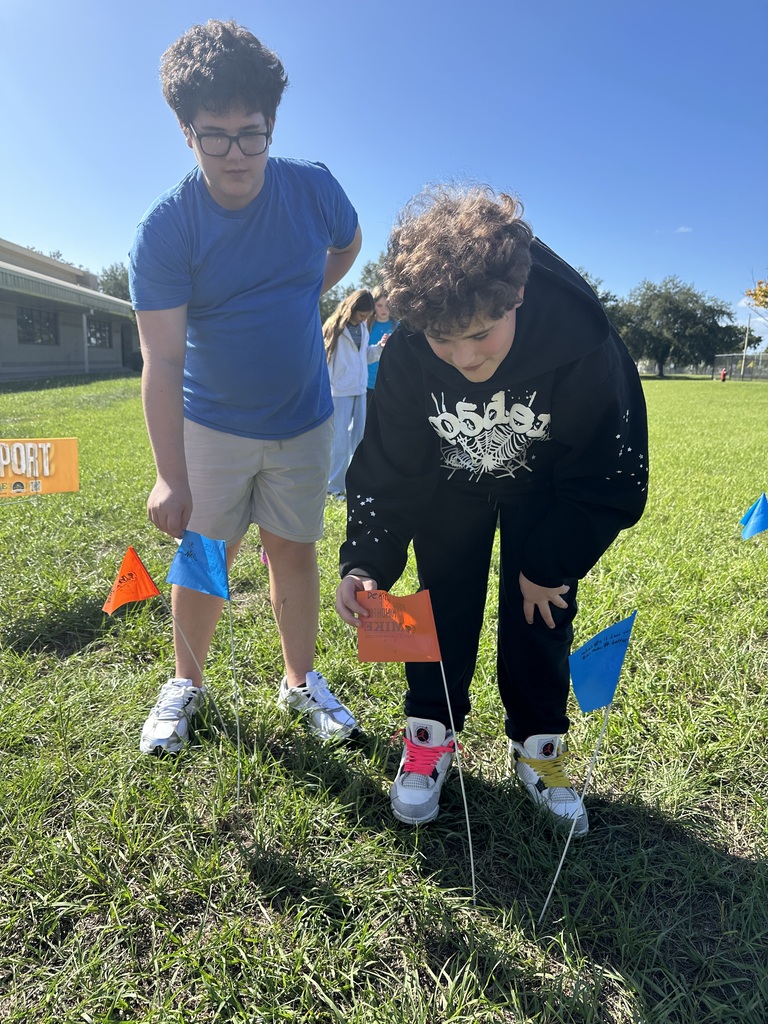 Two students in a field with orange flags. One student picks up a flag, while the other looks on.