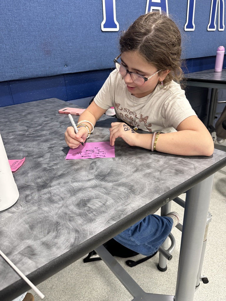 A student sits at a desk, writing on pink paper with a white marker.