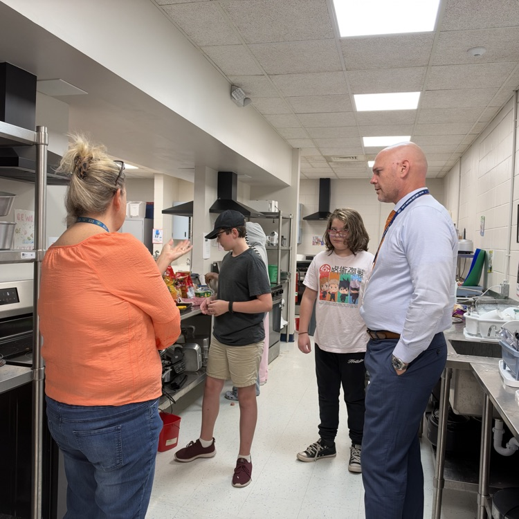 Four adults stand in a commercial kitchen with white walls and tiled floors. One holds a clipboard.