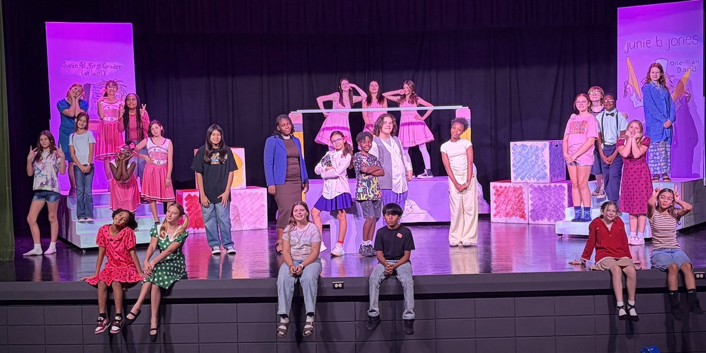 A group of students on stage with pink and purple stage props; some standing and some sitting