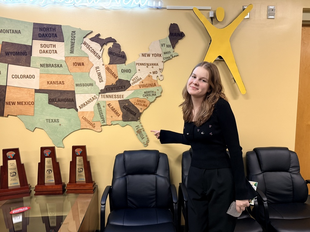 Woman in black suit holding paper stands in front of wall with map of USA and trophies.
