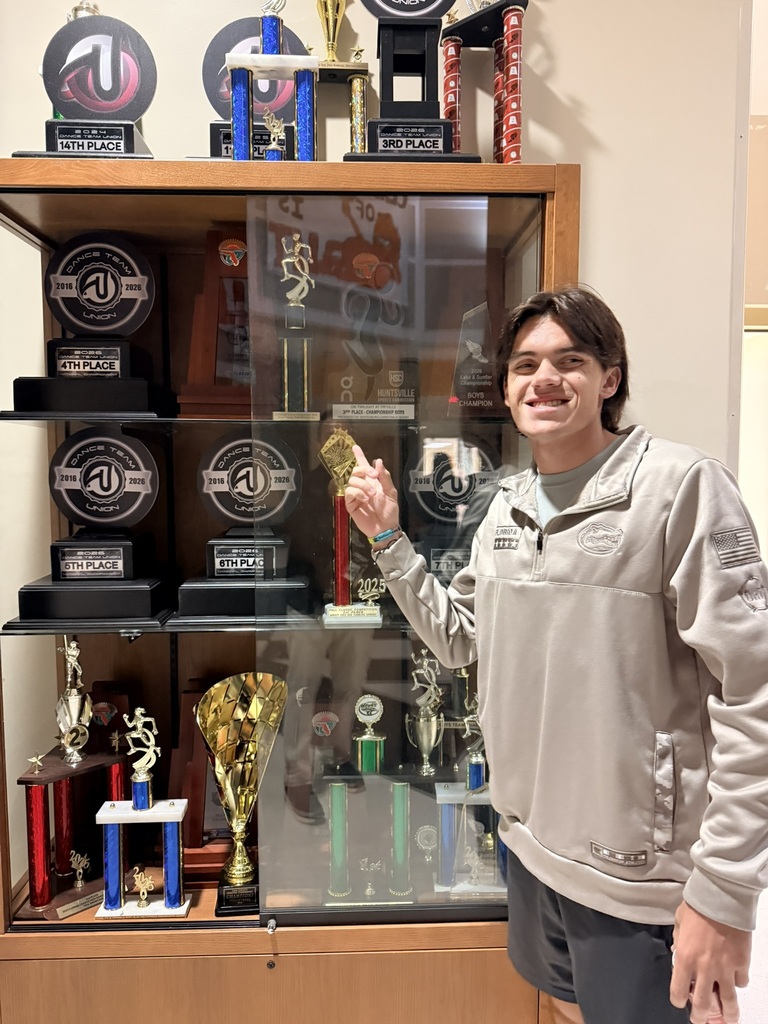 A man stands next to a cabinet with trophies. The cabinet has glass doors. He holds a trophy in his hand.