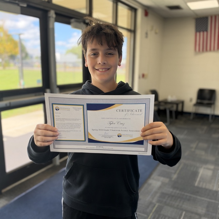 A student holds a certificate in both hands, smiling. Behind them is a glass door and a flag.
