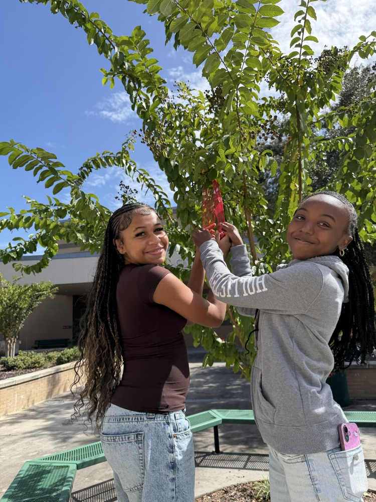 Two students in school uniforms hold a red ribbon tied to a tree outdoors.