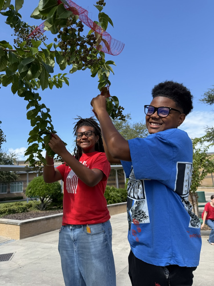 Two students hold up a branch with red berries and a ribbon outdoors. Behind them, a school building and landscaping are visible.