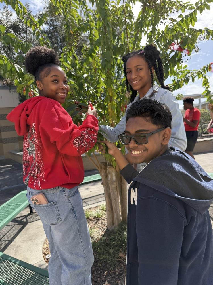 Three students stand outdoors, two girls and one boy, smiling near a small tree. One girl holds a branch.