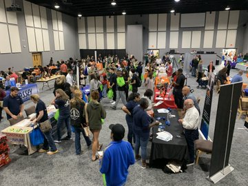 Many people at a career fair with tables and signs, one person speaking with another.