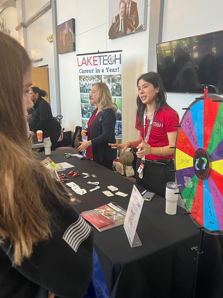 Two women in red shirts stand by a table with a colorful wheel, a stuffed animal, papers, and cups.