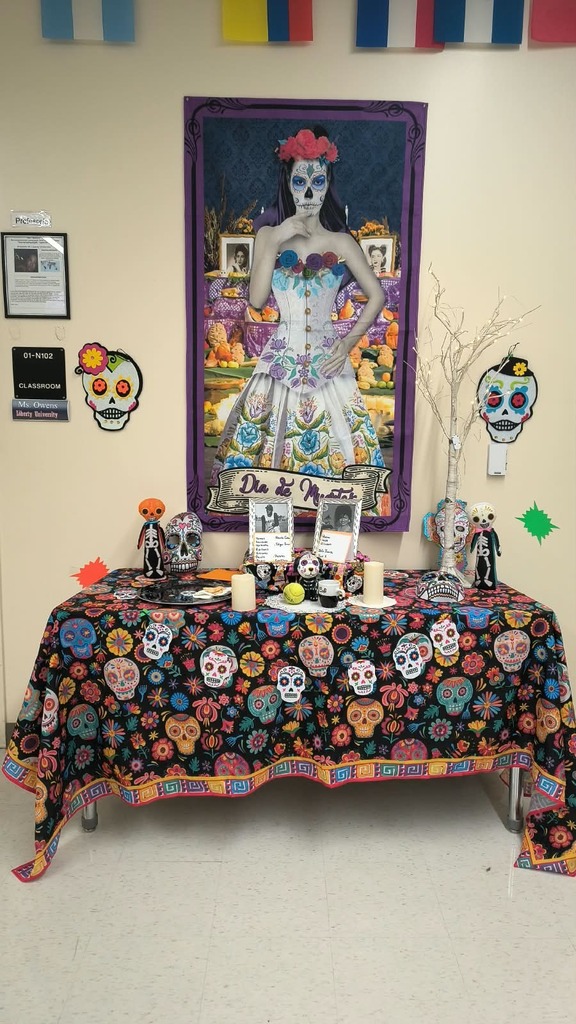 A table displays colorful skulls, candles, and decorative items. A framed painting of a woman in a floral dress is on the wall.