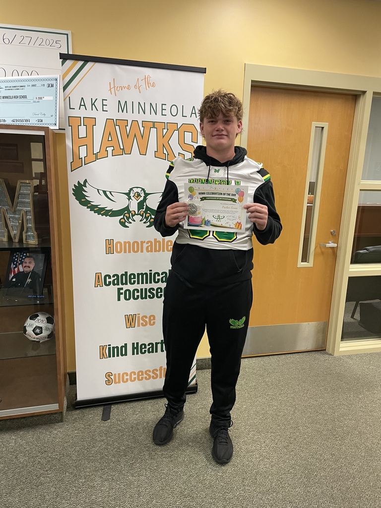 A student holds a certificate against a wall displaying a Hawks banner, a soccer ball, and a bookshelf.
