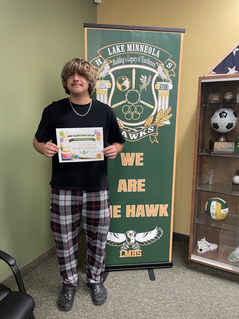A boy in plaid pants and a black T-shirt holds a certificate, standing in front of a banner.
