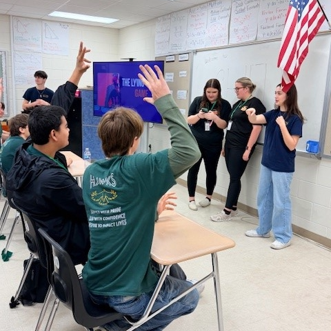 Students seated and raising their hands while other students stand at front of classroom and present