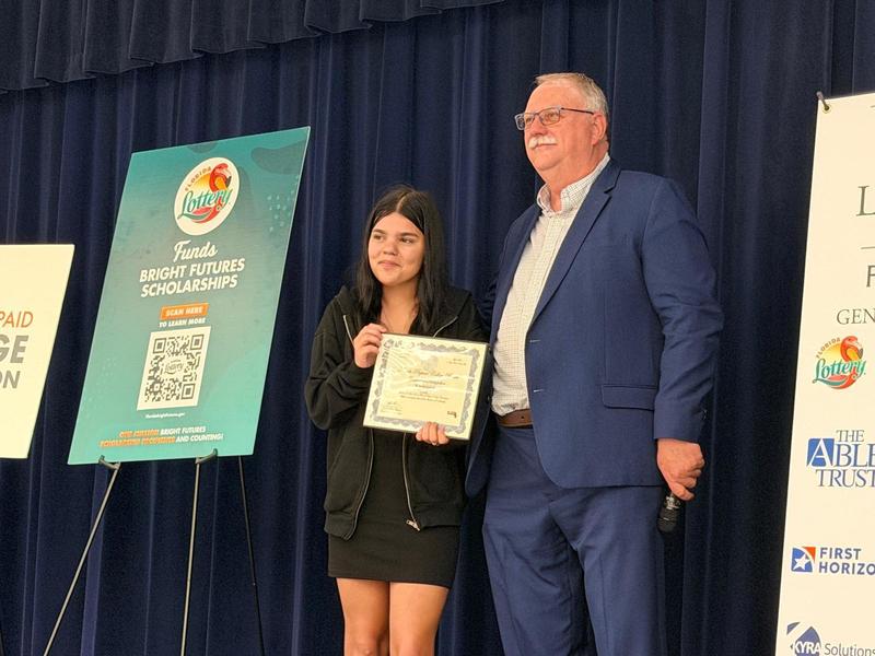Wes Lock standing on stage with student Millie Olvera, who is holding a plaque