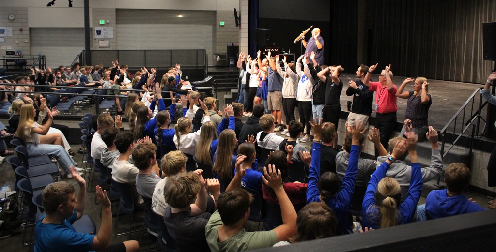 Teachers standing along the front edge of the stage and students raising hands mimicking teachers to one of the songs he played. 