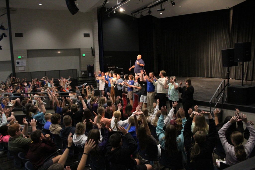 Reggie Dabbs speaking to teachers at the front of a stage with students listening in the auditorium.  