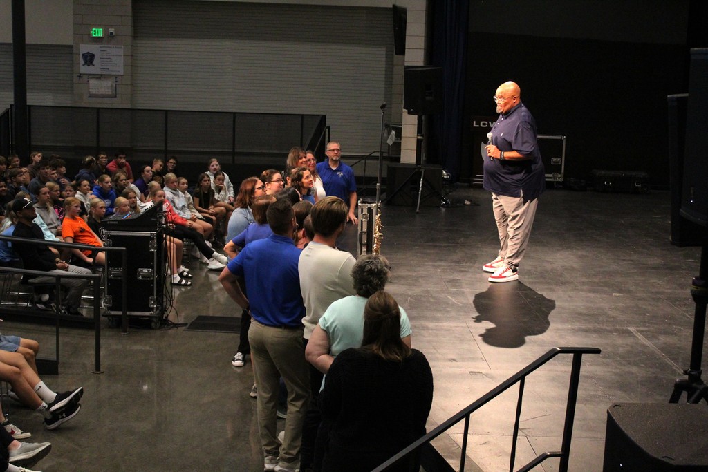 Reggie Dabbs speaking to teachers at the front of a stage with students listening in the auditorium.  