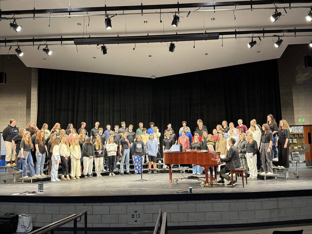 Both choirs standing on risers on the stage doing warm ups. 